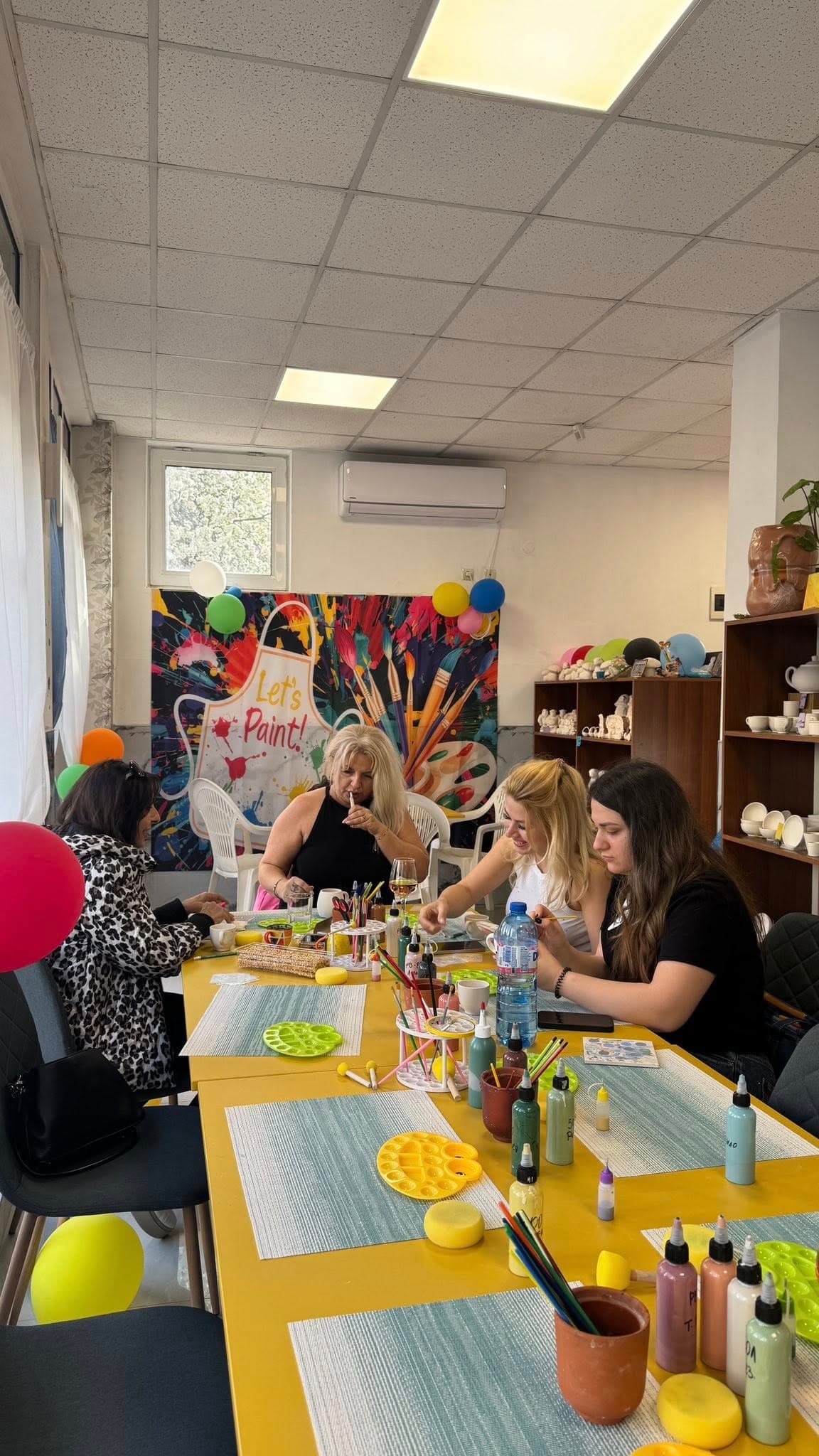Two friends laughing and painting ceramic mugs in a warm, sunlit studio
